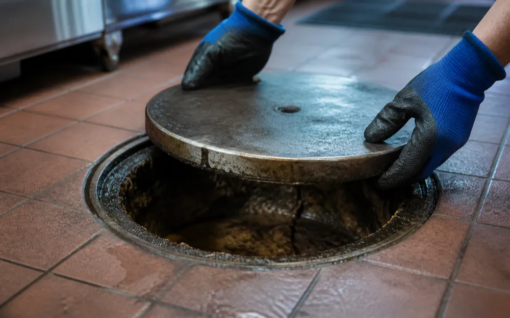 Gloved hands lifting the steel lid off a commercial kitchen grease interceptor set into a tiled floor