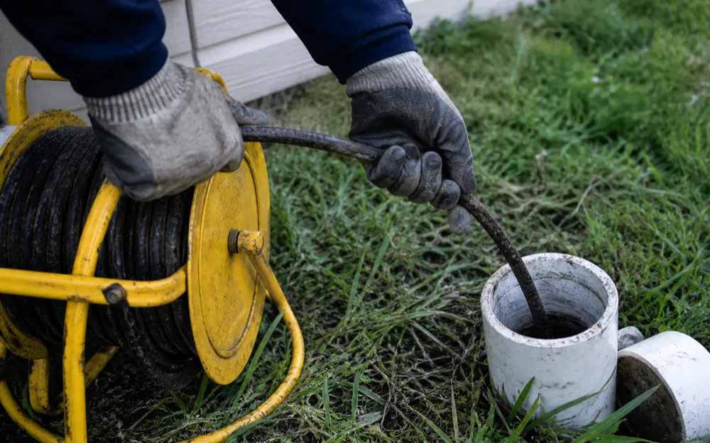 Gloved hands feeding a sewer inspection push-cable off its reel into an open cleanout in a side yard