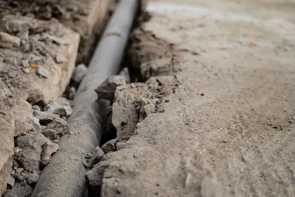 An exposed sewer line running along the bottom of an open excavation trench beside a concrete slab