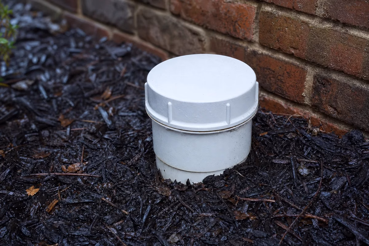 A white PVC sewer cleanout cap rising from a mulched bed beside the brick foundation of an older home after rain